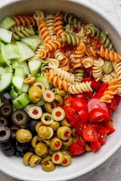 a white bowl filled with pasta, olives, tomatoes and cucumber slices