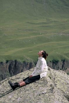 a woman sitting on top of a large rock next to a lush green hillside and valley