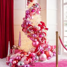 a christmas tree decorated with pink and red ornaments in front of a large window on a pink carpeted floor