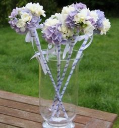 purple and white flowers in a glass vase on a wooden table with grass behind it