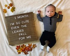 a baby laying next to a sign that says i month, i'm so cute even the leaves fall for me