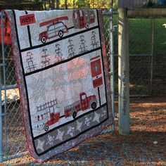 a quilt hanging on a chain link fence in front of a gate with cars and trucks