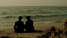 two people are sitting on the beach looking out at the water and sand in front of them