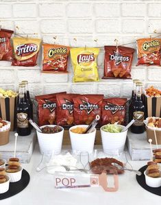 an assortment of snacks are displayed on a table in front of a brick wall with bags of chips and hotdogs hanging from it