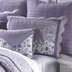 a bed topped with purple pillows next to a white nightstand and night stand in front of a window