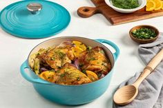 a blue pot filled with chicken and vegetables on top of a white table next to two wooden spoons