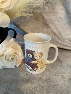 there is a coffee cup with two teddy bears on it next to some white flowers