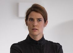 a woman wearing a black jacket is standing in front of a white wall and looking at the camera