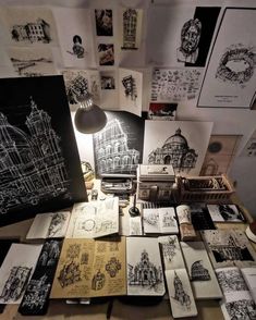 a table topped with lots of different types of drawings and pictures on paper next to a lamp