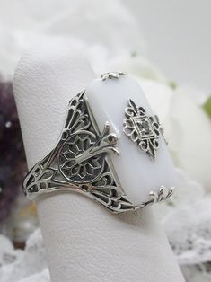 White Camphor Glass Solid Sterling Silver Ring Grace Design#D233 This is a lovely antique Edwardian era inspired filigree ring. This gorgeous ring is created in stunning white camphor glass. The glass is embellished with a delicate floral filigree. The top of the ring holds a center set 1mm round cut gemstone (Choose from white Cubic Zirconia, lab created white Moissanite, or Natural/Genuine White Diamond). The white glass is 18mm x 19mm. The ring sits 18mm north-south and 10mm east-west on the Camphor Glass Jewelry, Floral Filigree, Pink Snow, Edwardian Style, Gem Diamonds, Gorgeous Ring, North South, Filigree Design, Green Diamond