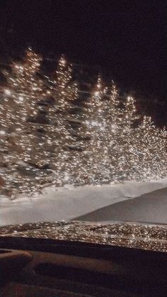 snow covered trees in the distance with lights on them at night, seen from inside a vehicle