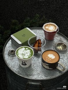 two cups of coffee sit on top of a metal table with other drinks and pastries