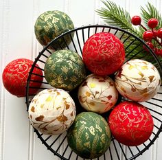 a basket filled with red and green balls next to a pine branch on top of a white wall