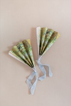 two paper fans with blue ribbons tied to them on a beige surface, one is folded and the other has an image of trees