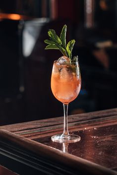 a drink in a wine glass with a green sprig on the top sits on a bar