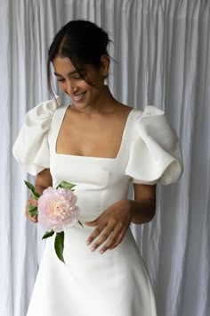 a woman in a white dress is holding a pink flower and smiling at the camera