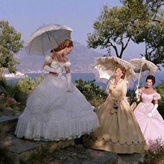 three women dressed in dresses and holding umbrellas on a hill overlooking the water with trees behind them