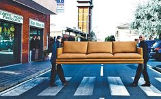 two men walking down the street carrying a couch across the street in front of them