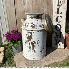 an old milk can sitting next to a welcome sign and potted plants on the ground