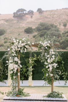 an outdoor ceremony setup with white flowers and greenery on wooden poles, in front of hedges