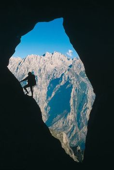 a man climbing up the side of a mountain in front of a cave with mountains behind him