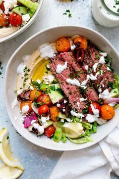 a white bowl filled with meat, tomatoes and lettuce next to a lemon wedge