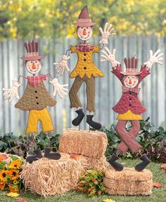 three scarecrows standing on hay bales in front of a fence and flowers