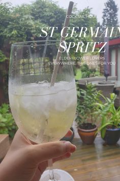 a person holding a wine glass with ice in it on a wooden table outside near potted plants