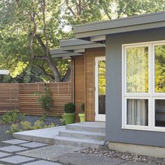 a small gray house sitting next to a lush green tree and shrubbery on the side of it