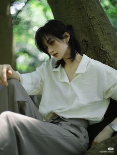 a woman sitting on the ground in front of a tree wearing a white shirt and gray pants