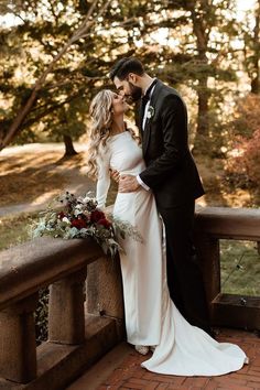 a bride and groom standing on a bridge in front of some trees with their arms around each other