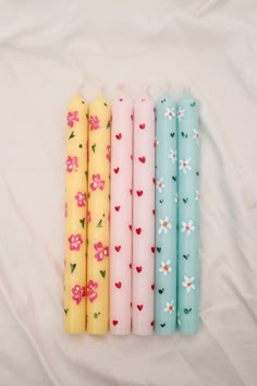 four different colored candles are lined up on a white sheet with pink, blue, and yellow flowers