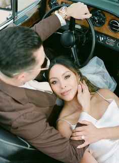 a bride and groom sitting in the driver's seat of a car, posing for a photo