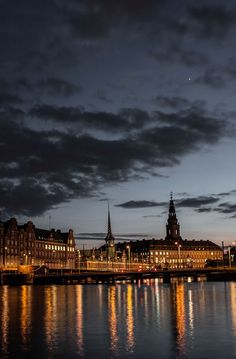 the city is lit up at night with lights reflecting in the water and dark clouds