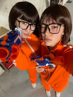 two girls in orange sweatshirts are holding remote controls and looking at the camera while standing next to each other