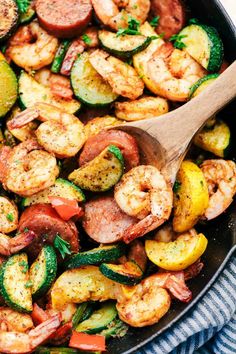 a skillet filled with shrimp, zucchini and vegetables next to a wooden spoon