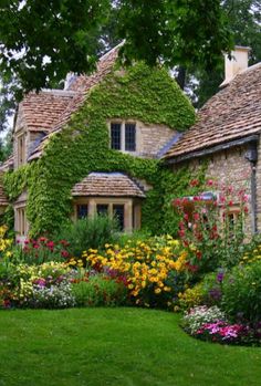 a house with flowers in front of it on the grass and trees around it, surrounded by greenery