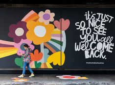 a woman walking past a wall with flowers painted on it and the words, it's just so nice to see you are welcome back