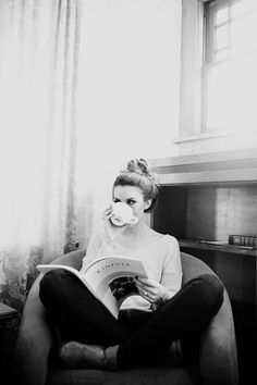 a woman sitting in a chair drinking from a cup and reading a book while holding a coffee mug