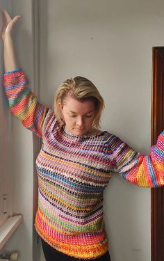 a woman leaning against a wall with her arms in the air, wearing a multicolored sweater