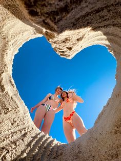 two women in bikinis standing on the beach looking out from behind a heart shaped hole