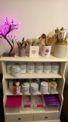 a white shelf filled with lots of paint and craft supplies next to a pink light