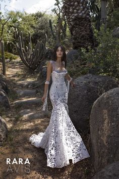 a woman in a white dress standing next to some rocks and cactus trees with her hands on her hips