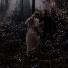 two people walking through the woods in front of some foggy trees and bushes, with one person wearing a long white dress