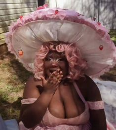 a woman with pink hair wearing a large hat and dress sitting on the ground in front of a house