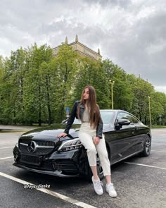 a woman sitting on the hood of a black car in a parking lot next to trees