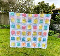 a crocheted blanket hanging on a clothes line in the grass with trees and bushes behind it