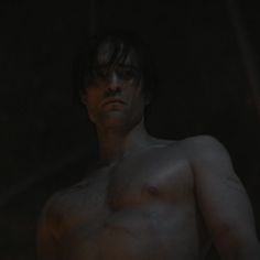 a shirtless man standing in the dark with his hand on his hip and looking off to the side