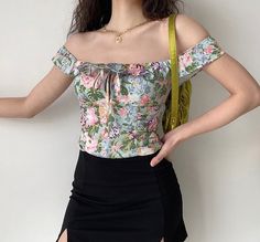 a woman wearing a floral top and black skirt with her hands on her hips, posing in front of a white wall