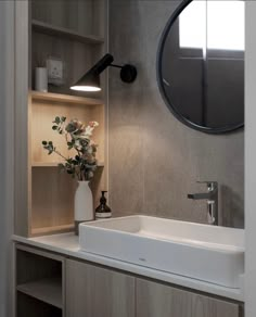 a white sink sitting under a round mirror next to a wooden shelf filled with flowers